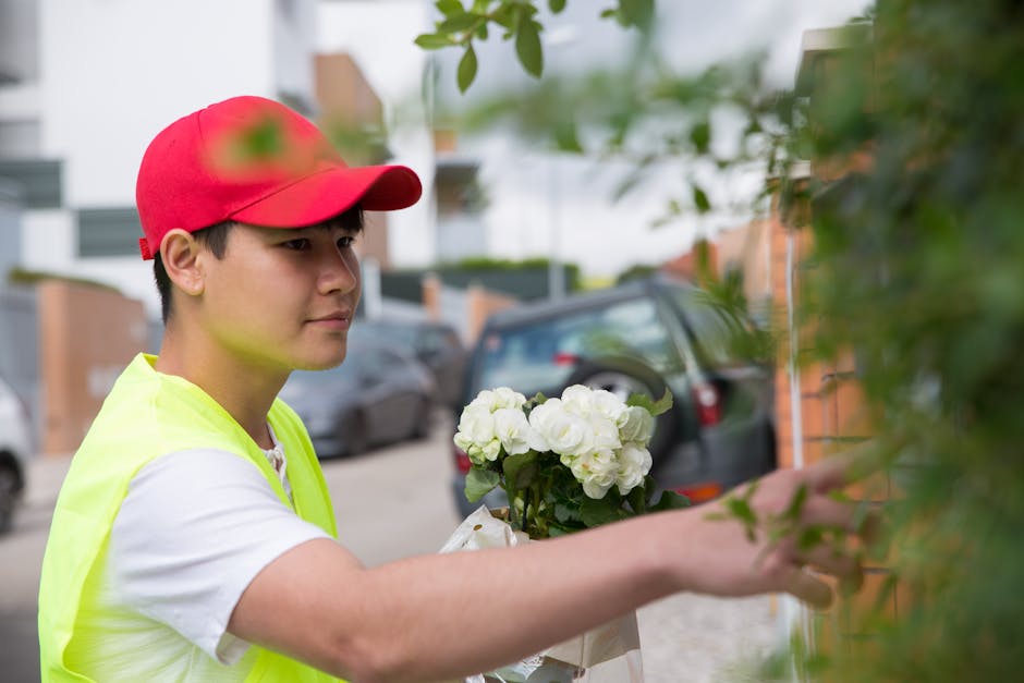 A young delivery person wearing a red cap and a neon yellow vest is placing a bouquet of fresh white flowers, including roses and hydrangeas, against a green leafy shrub outside a residential building