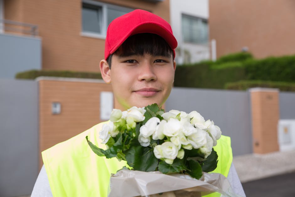 A young male flower delivery courier wearing a bright yellow high-visibility vest and a red cap holding a bouquet of fresh white begonias wrapped in white paper, standing outdoors in front of modern r