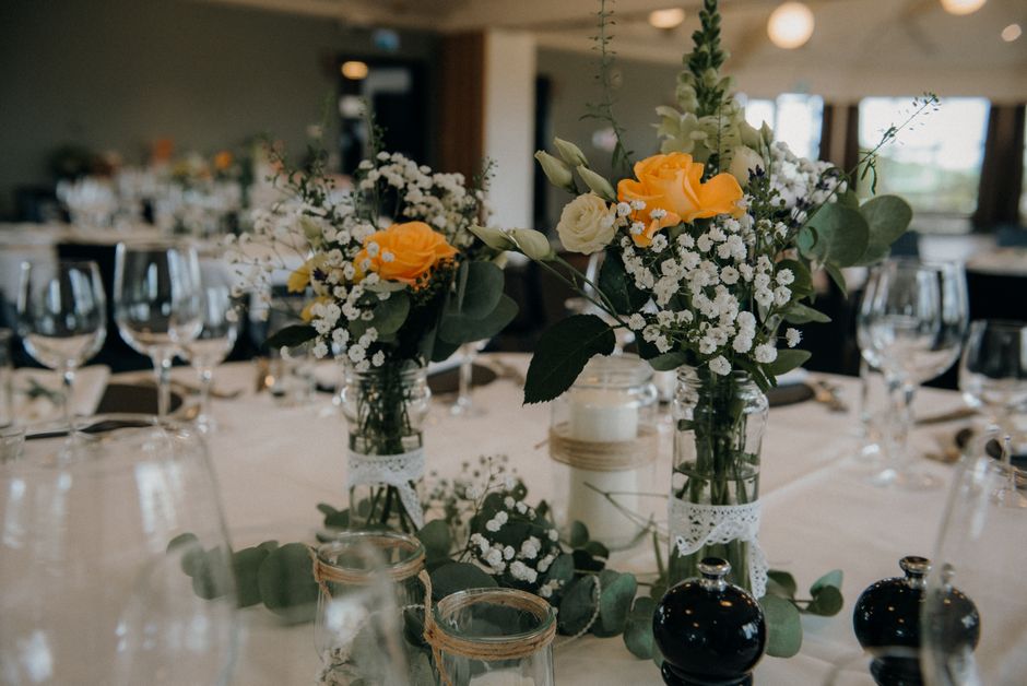 A floral arrangement featuring a mix of yellow roses, white lisianthus, and delicate white baby's breath, complemented by silvery-green eucalyptus leaves, situated in glass jars decorated with white l