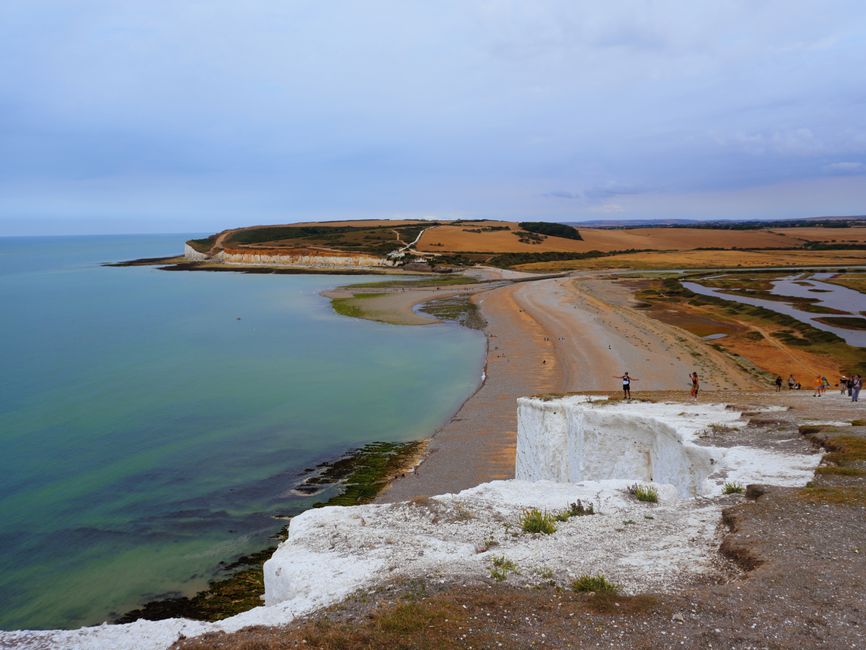 A scenic coastal landscape featuring a sandy beach with greenish water, bordered by white chalk cliffs. In the foreground, the edge of a white chalk cliff is visible, with a small patch of grass. Furt