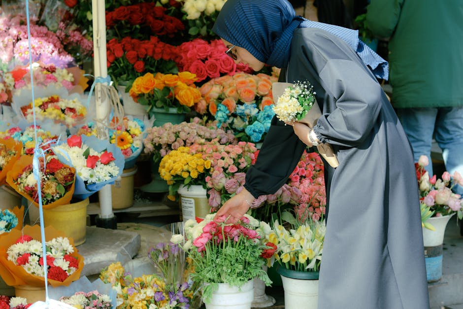 A woman wearing a blue hijab and a grey raincoat is selecting flowers at a busy outdoor flower market. She is holding a small bouquet of white and green flowers in her left hand while reaching out to 