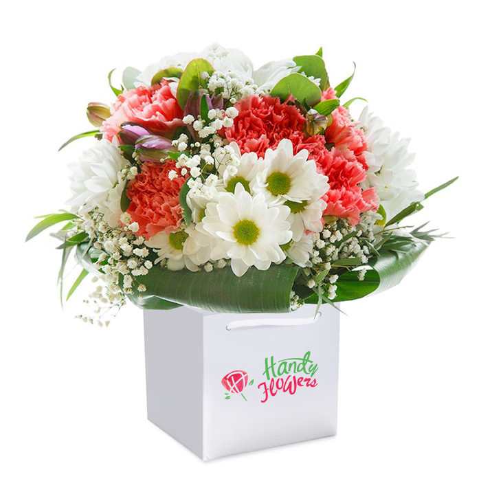 Resting upright in a clean-lined, white square gift bag, this harmonious arrangement is a testament to the expert craftsmanship found among Seven Sisters florists. The bouquet features plush coral and soft peach carnations, each blossom flaunting abundant ruffled petals that catch and reflect soft, natural light. Interspersed between these are radiant white daisies whose bold yellow-green centers add sparkle and contrast, reminiscent of springtime strolls through Downhills Park. The bouquet's lightness is enhanced by scattered sprays of fine baby's breath, providing a frothy, delicate counterpoint to the main flowers, while subtle buds with hints of purple and pink nestle at the edge to add interest and dimensionality. Everything is encircled by a thick wreath of broad, shiny green leaves, providing the display with depth and a finished, structured shape. White satin ribbon handles arch gracefully from the bag's top edge, promising easy, elegant gifting throughout Seven Sisters and the surrounding areas. Captured against a bright, seamless white background, every detail-the lush petals, the interplay of textures, the promise of unopened blooms-stands out in sharp relief. This bouquet is equally perfect for birthdays, anniversaries, or thoughtful surprises, its artistry and freshness conveying the essence of Seven Sisters' blossoming community with every stem.